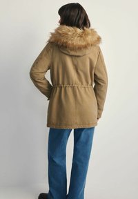 Brązowa parka z kapturem wykończonym futrem, z elastycznym paskiem w talii i długimi rękawami. Połączona z niebieskimi dżinsami o szerokich nogawkach oraz czarnymi botkami.