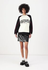 Felpa bianca e nera con motivo a scacchi e dettagli rossi, abbinata a una gonna in pelle nera con zip frontale. Scarpe chunky nere.