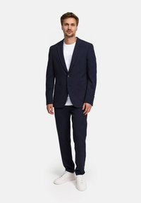 Abito blu navy con giacca a due bottoni e pantaloni coordinati, abbinato a una t-shirt bianca e sneakers bianche. Tessuto morbido e vestibilità moderna.