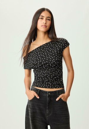 Schwarzes schulterfreies Oberteil mit weißen Punkten, das ein figurbetontes Design und eine geraffte Textur aufweist, kombiniert mit schwarzen High-Waisted-Jeans aus Denim.