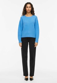 Pull bleu texturé avec un col rond, des manches longues et un ourlet côtelé, associé à un pantalon noir droit et des chaussures noires à bout pointu.