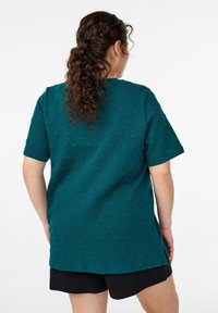 Petrolejové tričko s krátkým rukávem s texturovaným vzorem srdce, s volným střihem a bočními rozparky, nošené s černými šortkami.