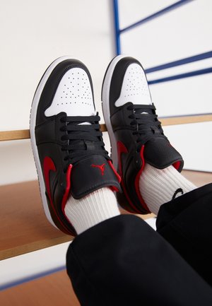 Stopy w czarno-biało-czerwonych butach Air Jordan z białymi prążkowanymi skarpetami, odpoczywające na drewnianych schodach z niebieską poręczą w tle.