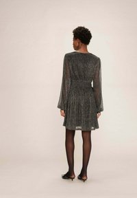 Una donna con i capelli corti e ricci è in piedi di spalle, indossando un vestito lungo a maniche lunghe di colore grigio scuro e luccicante, collant neri e décolleté neri con tacco a kitten.