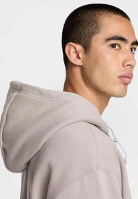 Hoodie gris clair en mélange de coton, avec capuche à cordon, larges poignets et coupe décontractée avec des détails de couture visibles.