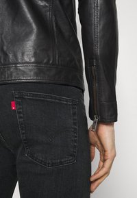 Närbild av en person med svart läderjacka med ärmzipp och svarta denimjeans med röd etikett på bakfickan.