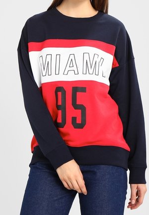 Vrouw draagt een marineblauwe, rode en witte trui met 'MIAMI' en '95' gedrukt op de voorkant, gecombineerd met een blauwe spijkerbroek.