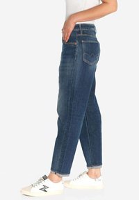 Donkerblauwe denim jeans met een tapered fit, voorzien van vijf zakken en zichtbare stiksels. Gecombineerd met witte sneakers.