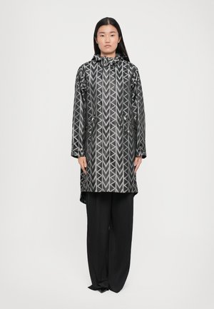 Cappotto impermeabile nero con un motivo geometrico argento, tasche con zip, cappuccio e vestibilità comoda. Abbinato a pantaloni neri larghi.