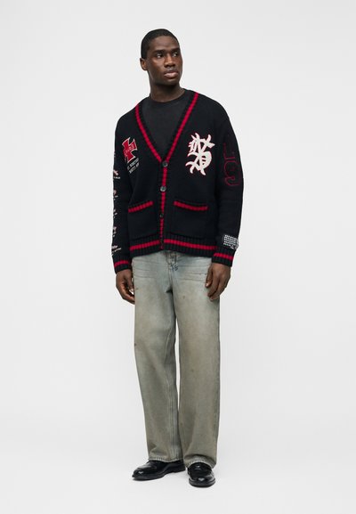 Άνδρας που φοράει μαύρη ζακέτα με κόκκινες λεπτομέρειες και μπαλώματα, μαύρο πουκάμισο, φαρδιά γκρι τζιν και μαύρα παπούτσια, στέκεται μπροστά σε λευκό φόντο.