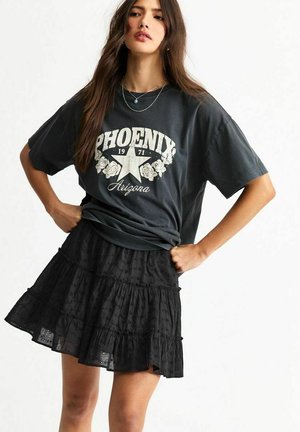 Camiseta gráfica en gris oscuro con la letra "Phoenix" y un diseño de estrella, combinada con una falda negra de capas con tejido texturizado.