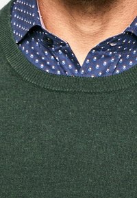 Pull vert en tricot avec un col rond, superposé sur une chemise bleu marine présentant des motifs contrastés à pois et rayures.