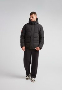 Mand iført sort pufferjakke med høj krave, sorte bukser og sort-hvide sneakers, stående foran en ensfarvet lys grå baggrund.