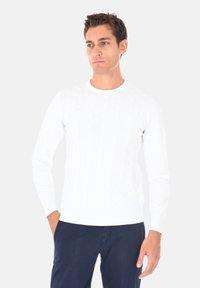 Maglione bianco a maglia con motivo a treccia, polsini a coste e scollatura rotonda, abbinato a pantaloni blu scuro.