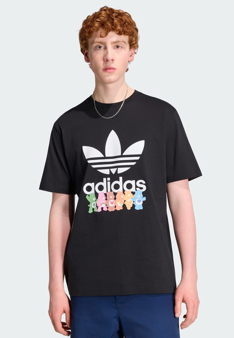 Junge Person mit lockigem roten Haar trägt ein schwarzes Adidas T-Shirt mit großem weißem Logo und bunten Care Bears, kombiniert mit blauer Hose.