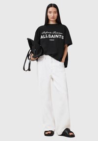 T-shirt nera oversize con stampa bianca che recita "Infinite Summer ALLSAINTS LONDON", abbinata a pantaloni bianchi a gamba larga e sandali neri.