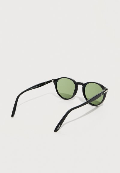 Μαύρα στρογγυλά γυαλιά ηλίου με πράσινους φακούς, λεπτά μπράτσα και διακριτικές ασημένιες λεπτομέρειες στους βραχίονες, τοποθετημένα σε λευκή επιφάνεια.