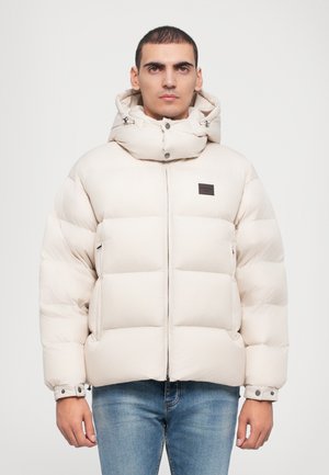 Chaqueta tipo plumón en color beige claro, con un alto cuello, cierre delantero y bolsillos laterales; construida con paneles acolchados para aislamiento.