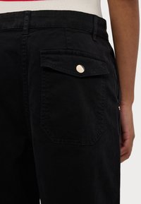 Pantalones negros de algodón con un acabado texturizado. Cuenta con un bolsillo trasero con una solapa redondeada, asegurada por un botón de presión metálico.