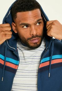 Navy zip-up hoodie with multicolored horizontal stripes (blue, orange, maroon) and a drawstring hood, layered over a striped shirt.