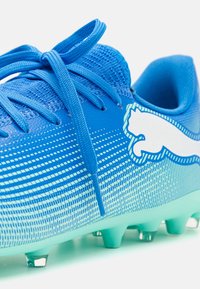 Bota de béisbol azul con parte superior de malla texturizada, diseño degradado, logo blanco en contraste y seis tacos verdes en la suela.