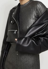 Veste courte en cuir noir avec fermetures éclair argentées, superposée sur une robe métallique foncée à côtes, mettant en valeur une texture lisse et un design structuré.