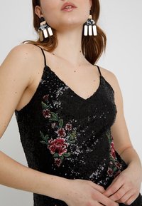 Černé šaty s flitrovou výšivkou a květinovými motivy; mají výstřih do V a tenká ramínka. Viditelné výrazné náušnice.