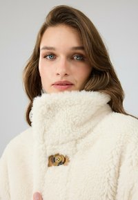 Crème kleurige, fluffy fleecejas met een hoge kraag en een ronde houten knoopsluiting. Zachte textuur en casual ontwerp.