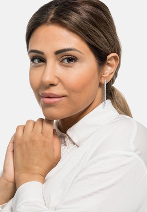 Orecchini a goccia lunghi in argento con cristalli rotondi trasparenti. Il modello indossa una camicia bianca con colletto, con le mani incrociate vicino al mento. Trucco naturale e gradevole.