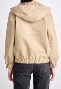 Beżowa kurtka z kapturem, z elastycznymi mankietami i paskiem w talii, wykonana z gładkiego materiału. Charakteryzuje się prostym wzornictwem z minimalnymi detalami na plecach.