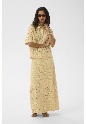 Femme portant un haut jaune en dentelle à manches courtes et une jupe longue assortie, des lunettes de soleil marron, un bracelet argenté et des sandales ouvertes, debout les bras croisés.