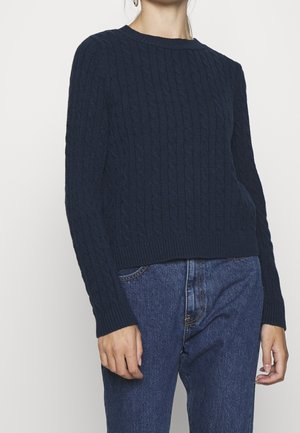 Frau trägt einen marineblauen, kabelgestrickten Pullover mit langen Ärmeln, kombiniert mit hoch-taillierten blauen Jeans vor einem schlichten Hintergrund.