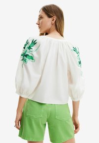 Blusa bianca con ricami floreali verdi sulle maniche, polsini a sbuffo e una vestibilità ampia. Abbinata a pantaloni corti in denim verdi.