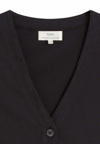 Primer plano del cuello de una camiseta negra con cuello en V, un botón y una etiqueta blanca que muestra "Yerse Organic Collection" y talla pequeña.