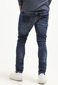 Jeans skinny de mezclilla azul oscuro con desgastes y dos bolsillos traseros. Llevados con zapatillas blancas sobre un fondo claro.