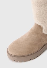 Beige suède winterboot met dikke rubberen zool en crèmekleurige sherpa voering zichtbaar bij de enkel.