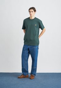 Tmavě zelené bavlněné tričko s krátkým rukávem a malým logem. V kombinaci s volnými modrými džínami a hnědými botami. Jednoduchý design, uvolněný střih.