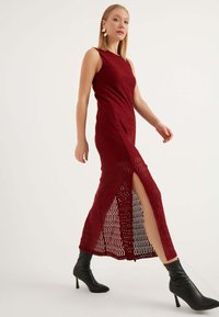 Maxi dress in magenta a maniche lunghe con pattern a zigzag, design senza maniche, spacco laterale, abbinato a stivali neri con tacco alto alla caviglia.