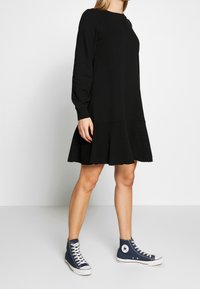 Vestido negro de manga larga con dobladillo acampanado, que presenta un escote redondo y una textura suave, combinado con zapatillas altas azules.