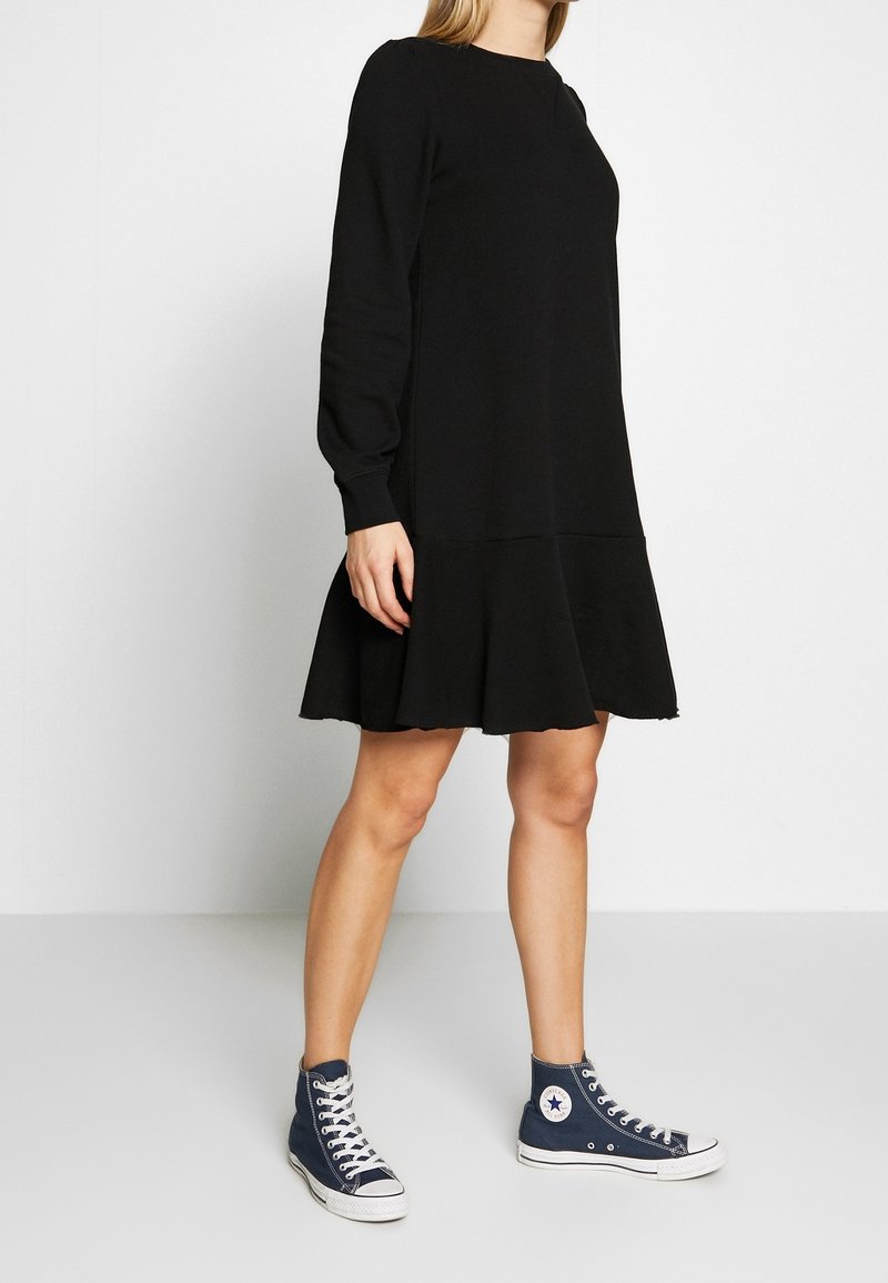 Vestido negro de manga larga con dobladillo acampanado, que presenta un escote redondo y una textura suave, combinado con zapatillas altas azules.