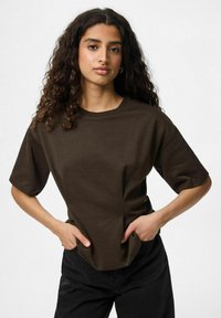 Brūns kokvilnas T-krekls ar īsām piedurknēm, brīvs griezums un apaļš kakla izgriezums. Modelis stāv ar rokām uz gurniem pret neitrālu fona.