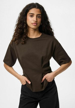 T-shirt en coton marron à manches courtes, coupe décontractée et col rond. Le mannequin se tient avec les mains sur les hanches contre un fond neutre.
