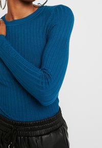 Pull en maille côtelée couleur sarcelle avec un col rond et des manches longues, assorti à un pantalon de jogging noir à taille élastique. Le tissu semble doux et extensible.