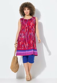 Bezramenné tunikové šaty v živé červené barvě s abstraktními vzory listů, kombinované s modrými capri kalhotami a béžovými sandály na klínu, držící tkanou tašku.