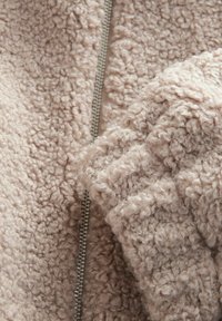 Lys beige, fluffy fleece stof med en tekstureret overflade; har en sølv lynlås langs sømmen og bløde, krøllede plysdetaljer.