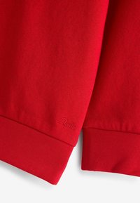 Pull en coton rouge avec des poignets et un ourlet côtelés. Présente un logo en relief sur le poignet gauche. Texture lisse et couleur uniforme sur l'ensemble.