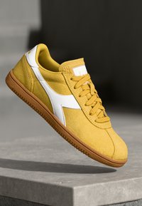 Yellow suede sneaker with a white leather side accent, round laces, and a brown rubber sole, featuring a padded collar and logo patch.