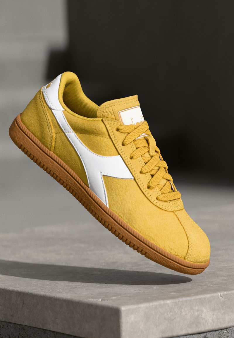 Yellow suede sneaker with a white leather side accent, round laces, and a brown rubber sole, featuring a padded collar and logo patch.