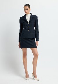 Blazer de rayas Navy con botones dorados, dos bolsillos; combinado con una falda navy ajustada; llevado con tacones altos color marfil. Textura de tela suave.