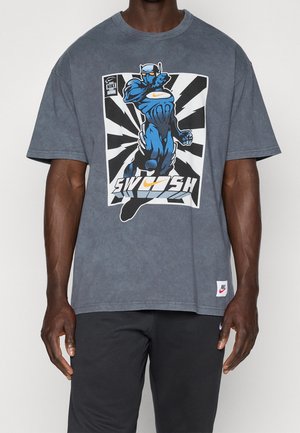 Personne portant un T-shirt Nike gris foncé avec un graphisme de super-héros bleu, un fond de rayons noirs et blancs, et le texte "SWOOSH" accompagné du logo Nike.